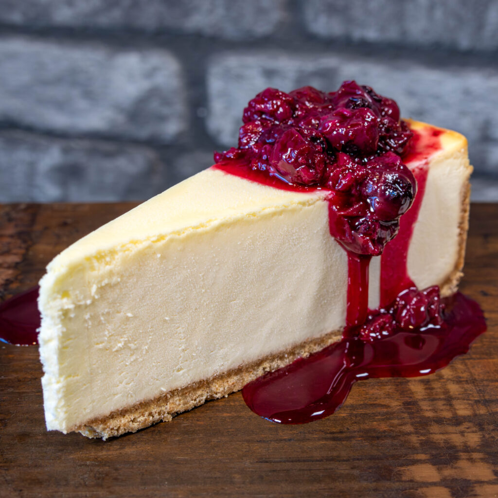 Slice of cheesecake with graham cracker crust and raspberry topping on wood serving board